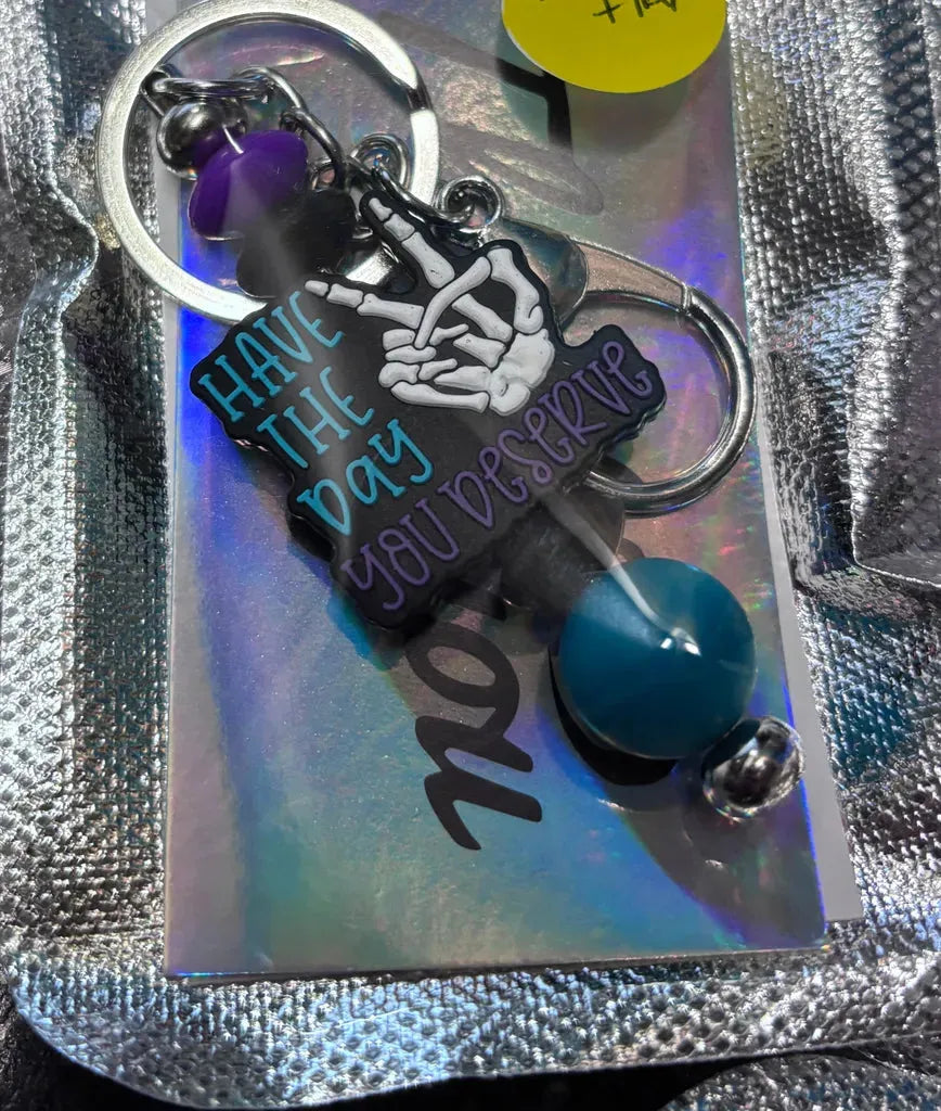 A custom beaded keychain with colorful beads and a charm that says "Have the Day You Deserve," displayed on iridescent packaging.